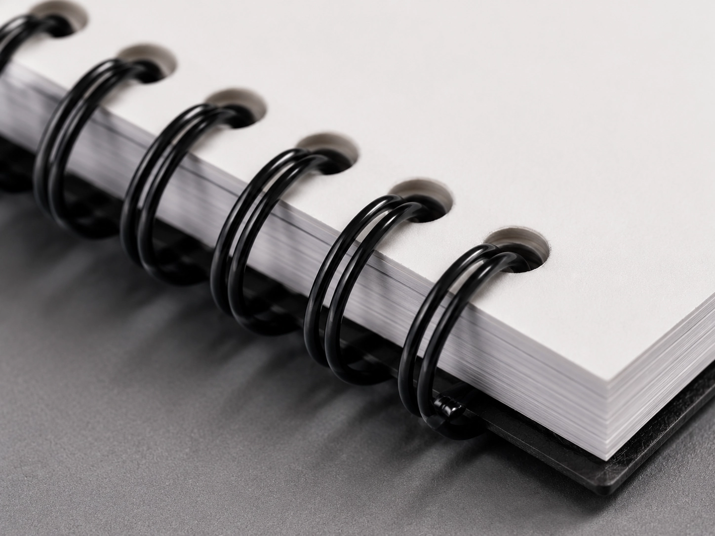 Macro close-up of a high-quality metal spiral single coil binding threaded through clean round punched holes of premium white paper, showing the professional crimped-off headed ends that prevent paper snags.